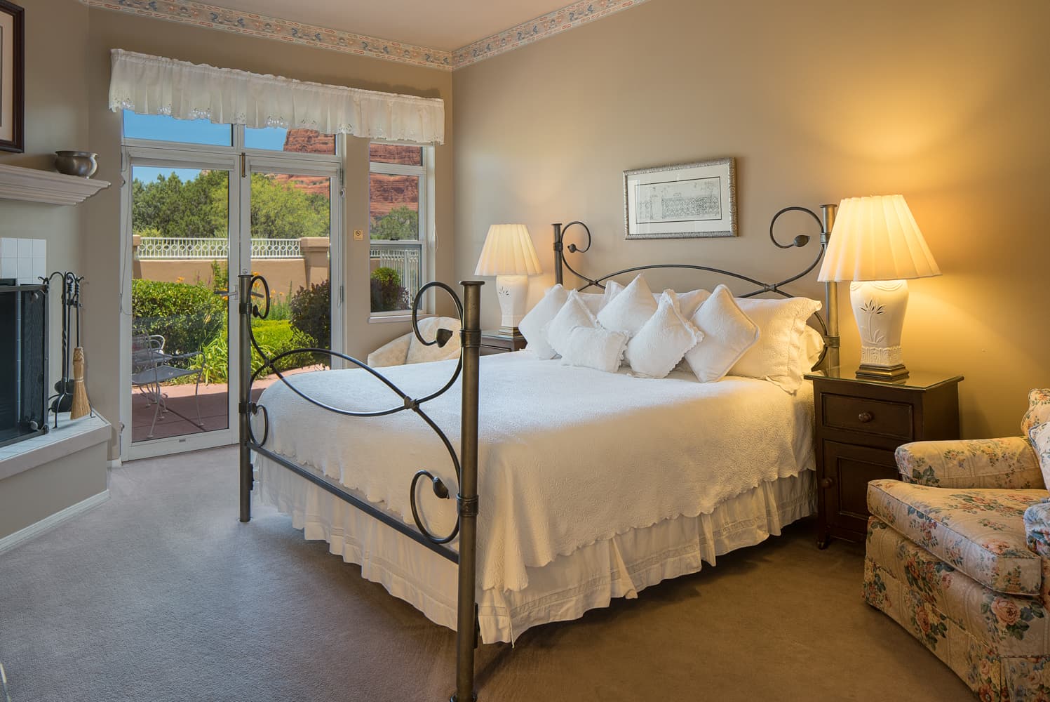 A well-appointed bedroom with a king-size bed, white bedding, two bedside lamps, and a fireplace, with a glass door opening to an outdoor patio.