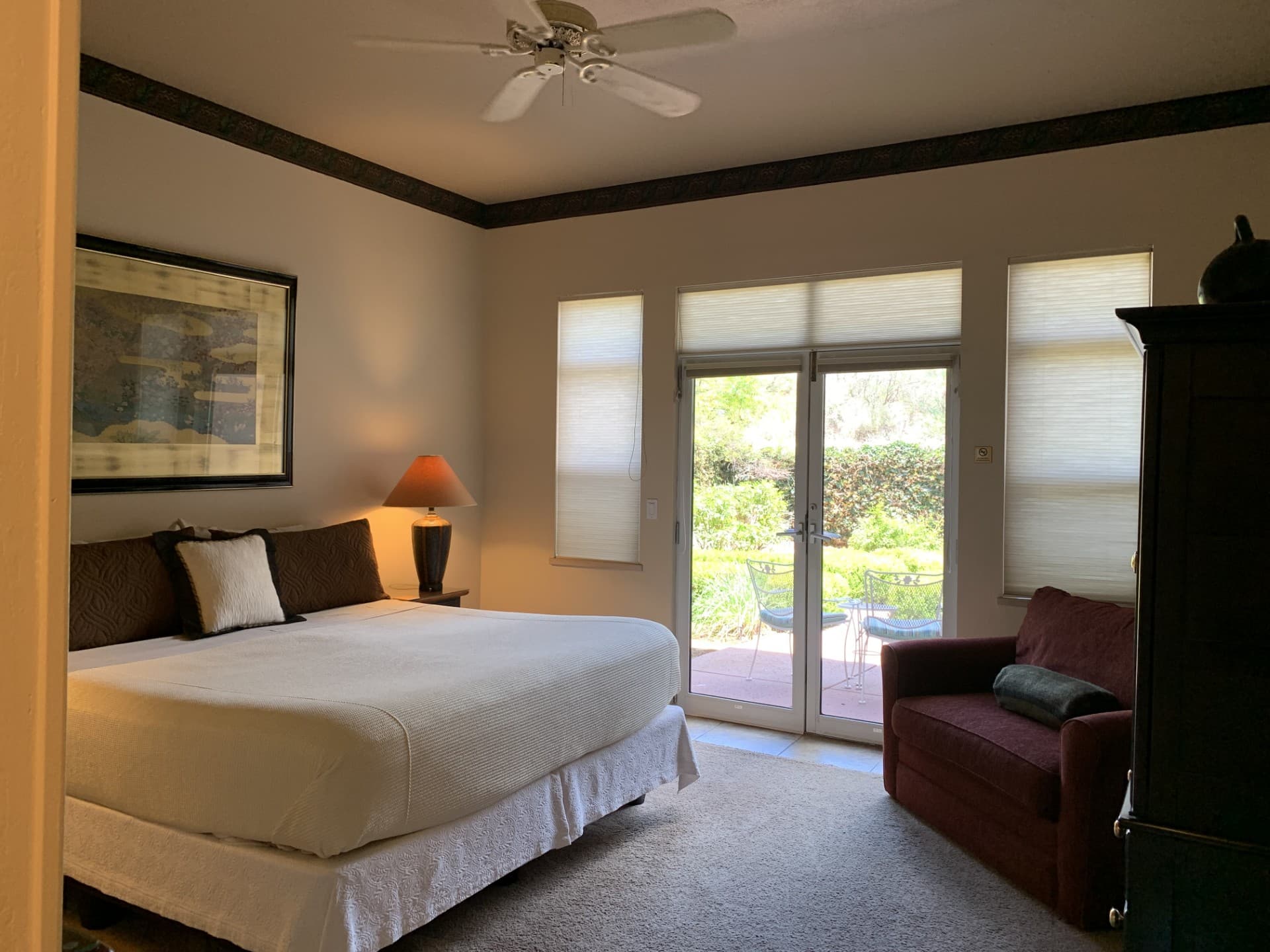 A well-appointed bedroom with a king-sized bed, two lamps, a red love seat, and large glass doors leading to an outdoor patio with garden views.