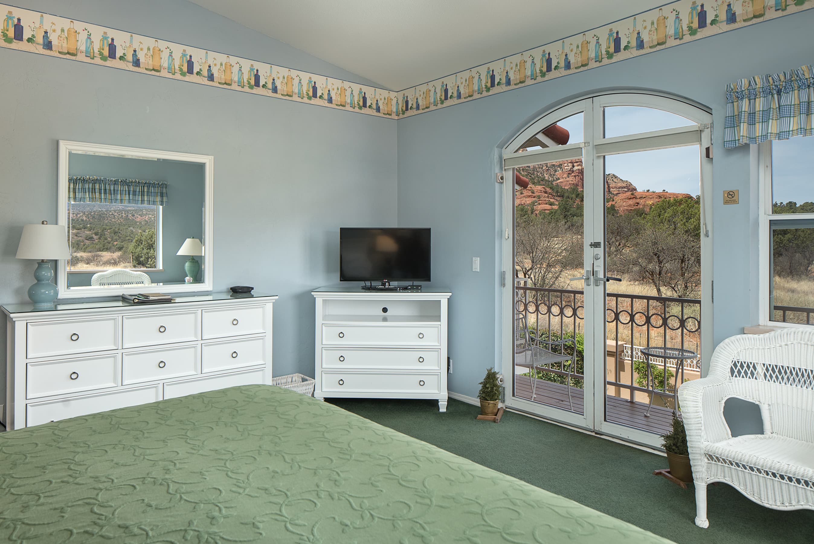 A bedroom with light blue walls, a green bedspread, white dressers, and a TV. An arched glass door opens to a balcony with views of red rock formations.