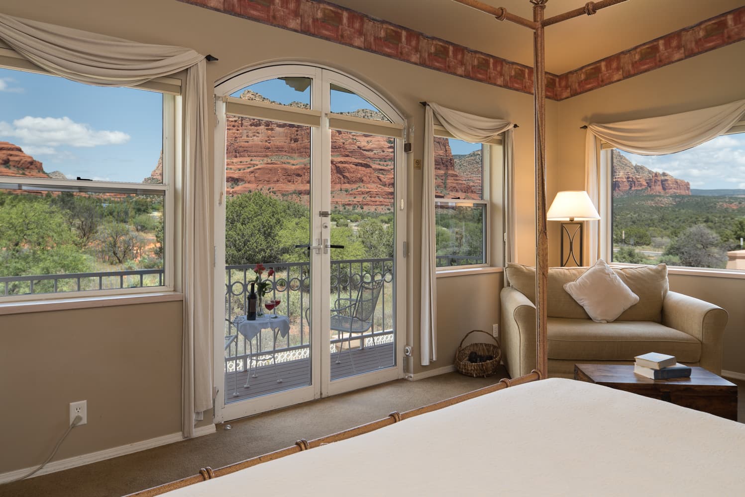 A charming room with a four-poster bed, cozy seating area, and a balcony with a table and wine, offering spectacular views of red rock formations.