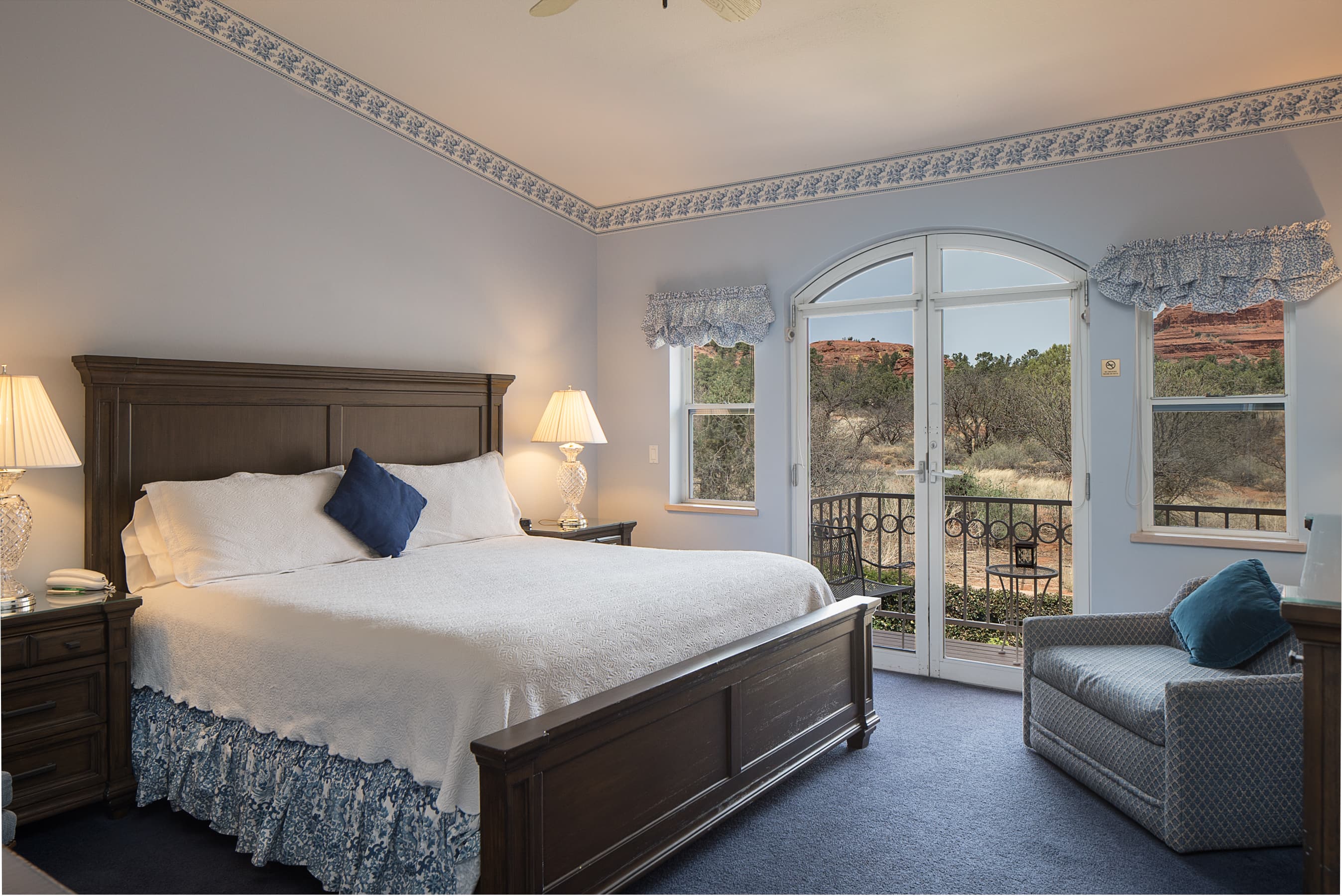 A serene bedroom with light blue walls, a dark wood king bed, and a cozy armchair. French doors open to a balcony with picturesque views of red rock formations.