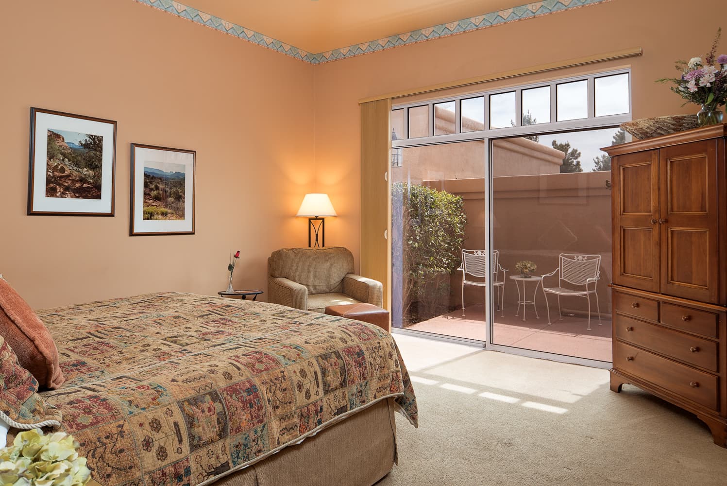 A warm-toned bedroom with a patterned bedspread, an armchair, and a wooden armoire. Sliding glass doors lead to a private outdoor patio with seating.