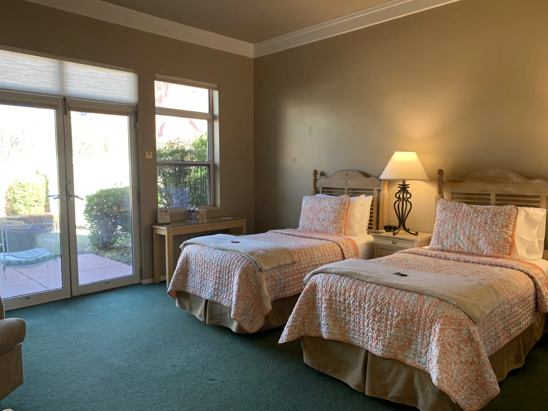 Sunny room with a 2 Twin bed, a wooden wardrobe, and an arm chair. French doors open to a patio with lush greenery and distant red rock formations.