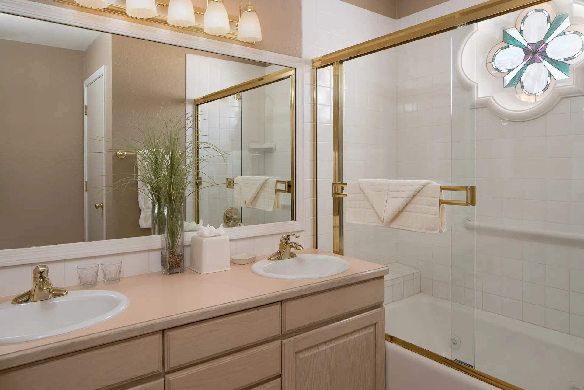 A well-lit bathroom with a double vanity, large mirror, and a combined bathtub and shower featuring a custom stained glass window.