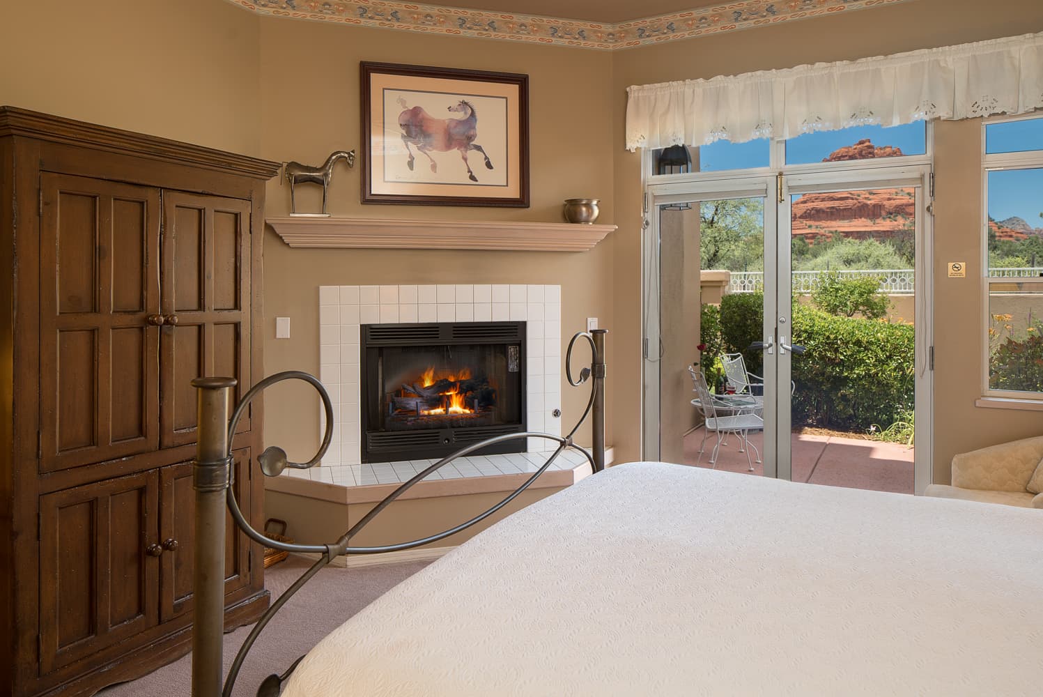 An inviting bedroom featuring a lit fireplace, a large wooden armoire, and a comfortable bed, with a glass door leading to an outdoor patio.