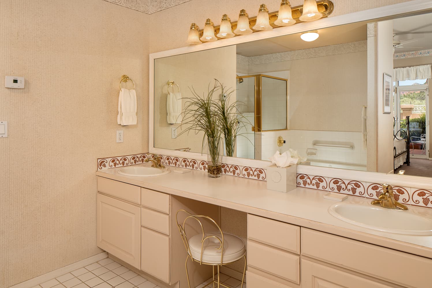 A spacious bathroom featuring a long vanity with two sinks, a large mirror, ample counter space, and a separate seating area under a bright light fixture.