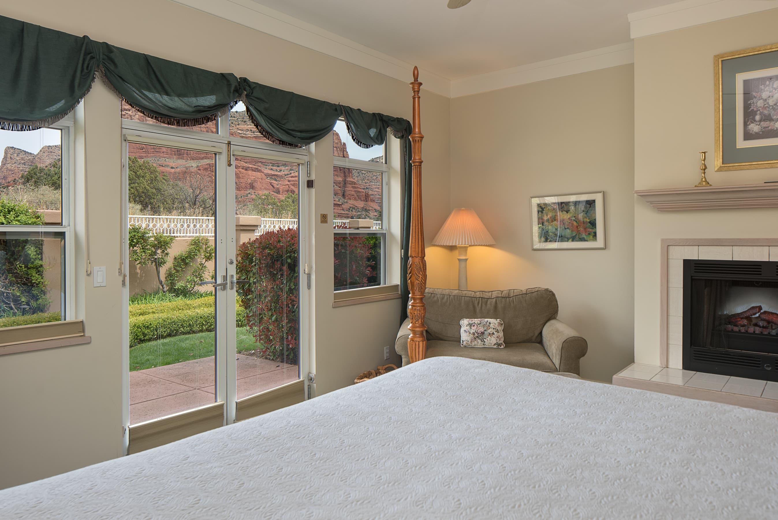 A bright bedroom with a bed, a cozy armchair, and a fireplace. French doors with dark green valances open to a patio with views of red rock formations.