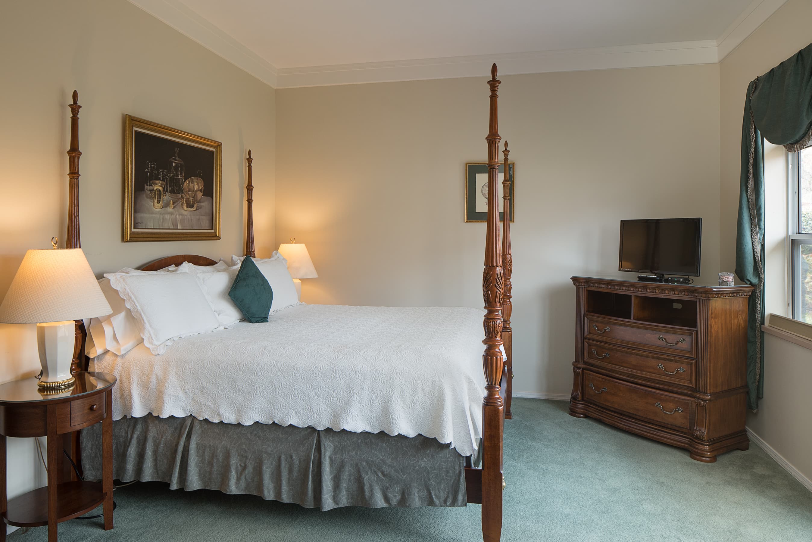 A spacious bedroom with a four-poster bed, white bedding, and two bedside tables with lamps. A dresser with a TV sits near a window with green drapes.