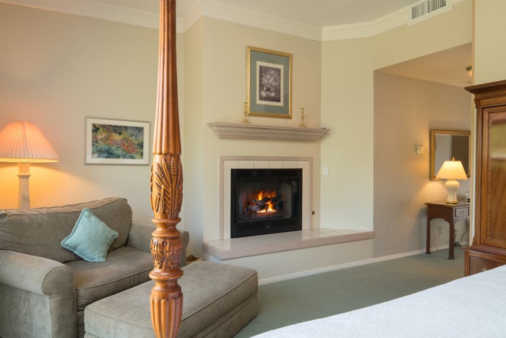 A cozy corner in a guest room with a lit fireplace, a comfortable armchair with an ottoman, and a side table with a lamp. A glimpse of a wooden four-poster bed is visible.