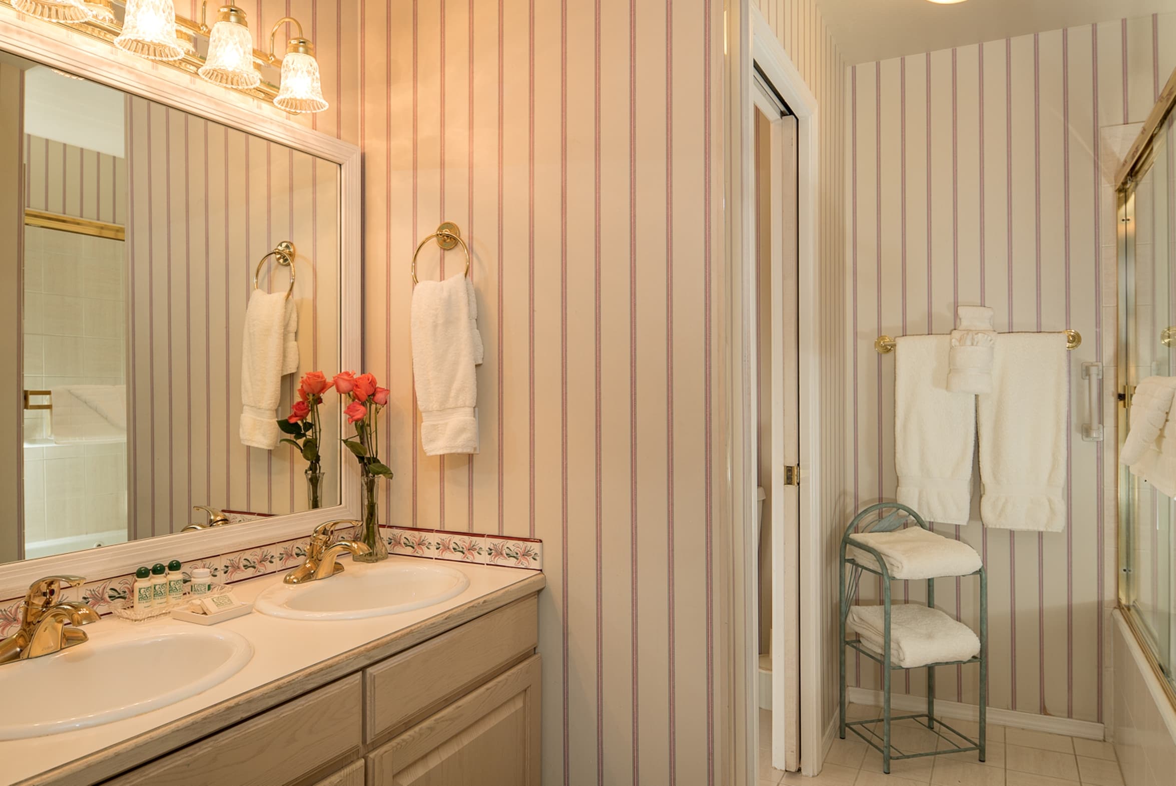 A brightly lit bathroom with striped wallpaper, a double vanity with two sinks, a large mirror, and a rack of fresh white towels.