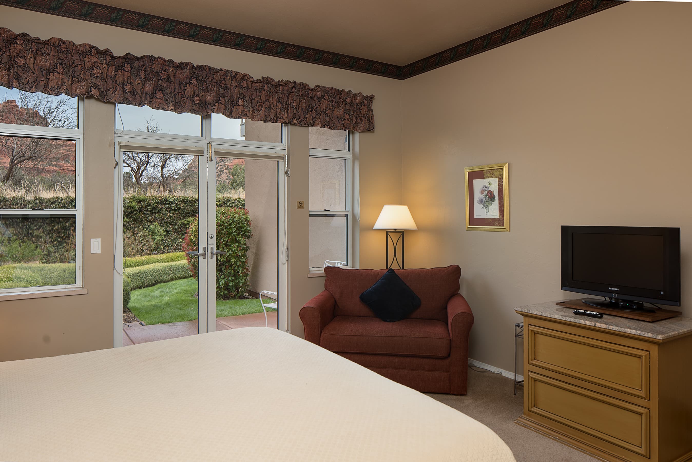 A spacious bedroom featuring a bed, a red love seat, a dresser with a TV, and glass doors opening to a lush outdoor patio.