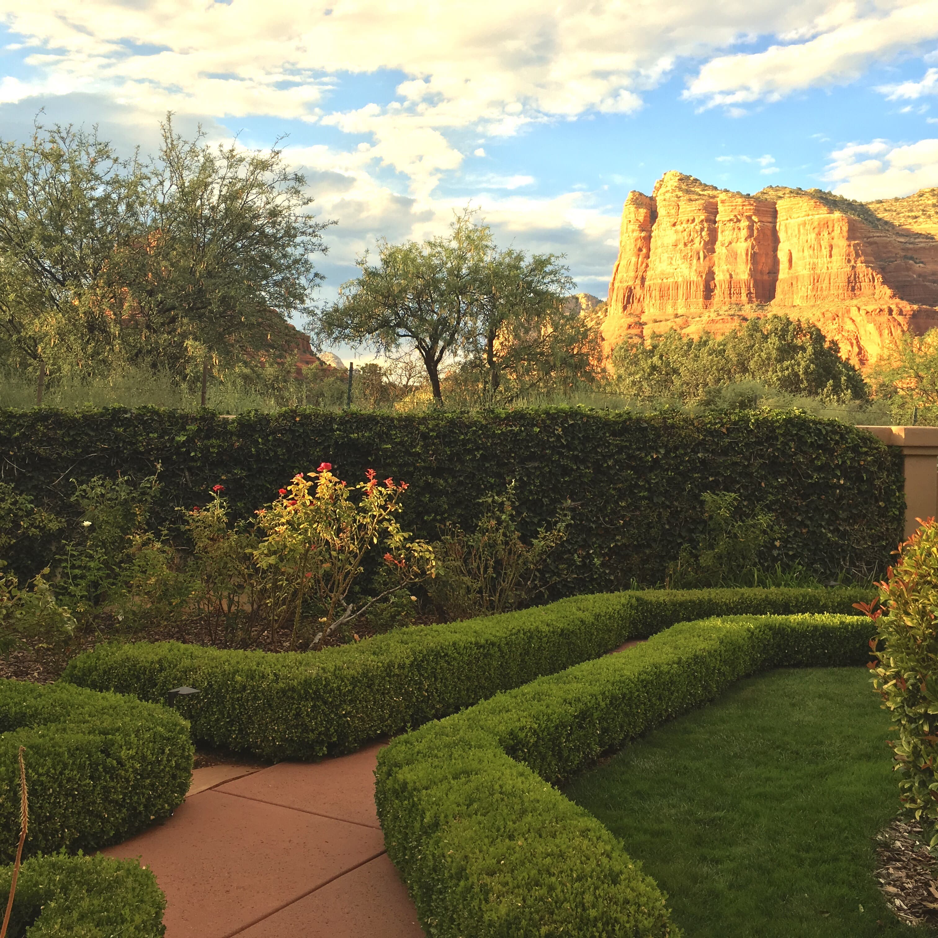 A vibrant garden path with manicured hedges and a lush lawn, set against a stunning backdrop of towering red rock formations under a partly cloudy sky.