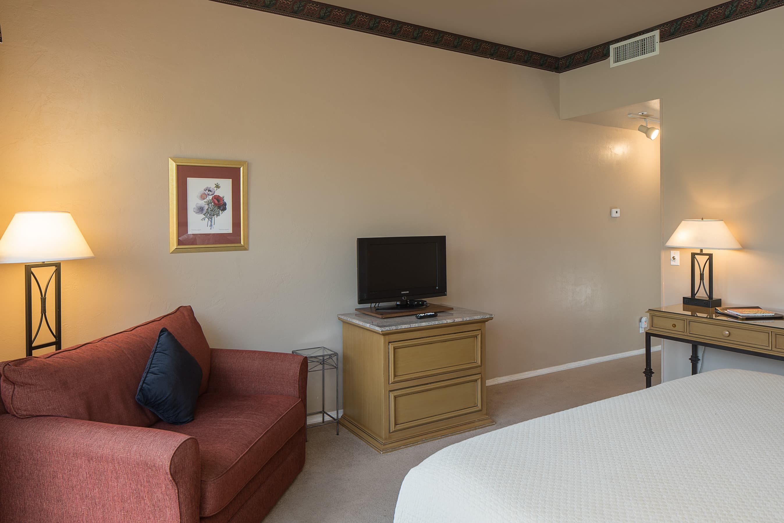 A guest room with a red armchair, a bedside lamp, a TV on a dresser, and a glimpse of a bed, all set against a neutral wall with decorative trim.