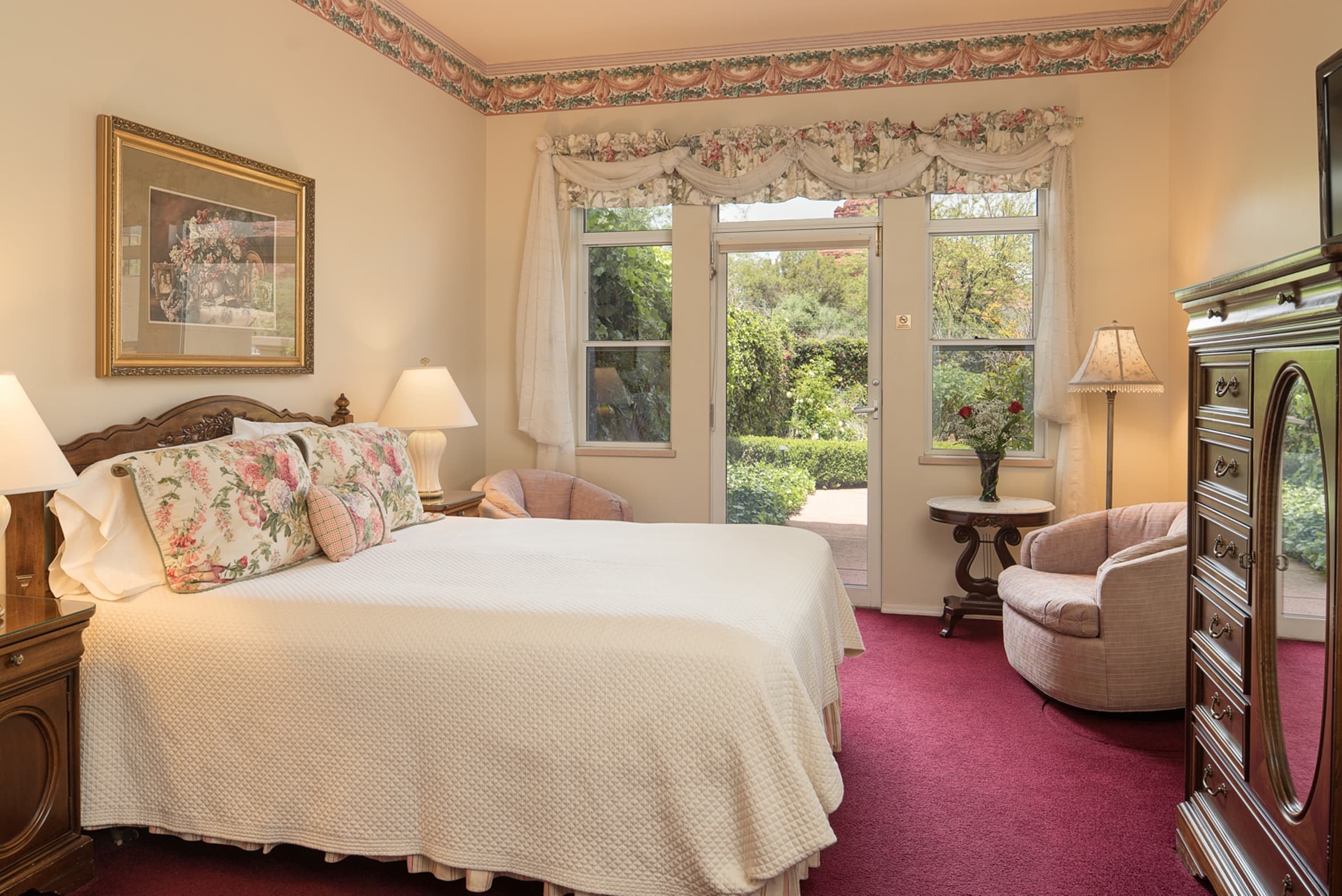 A spacious bedroom with a queen bed, floral accents, two armchairs, and a dresser. French doors open to a lush green outdoor area, with a vibrant red carpet.