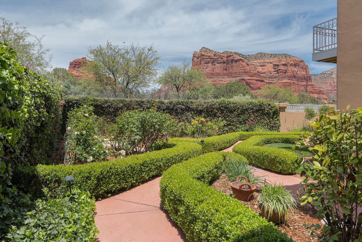 A vibrant garden with winding pathways and manicured hedges, set against the majestic backdrop of red rock mountains under a partly cloudy sky.