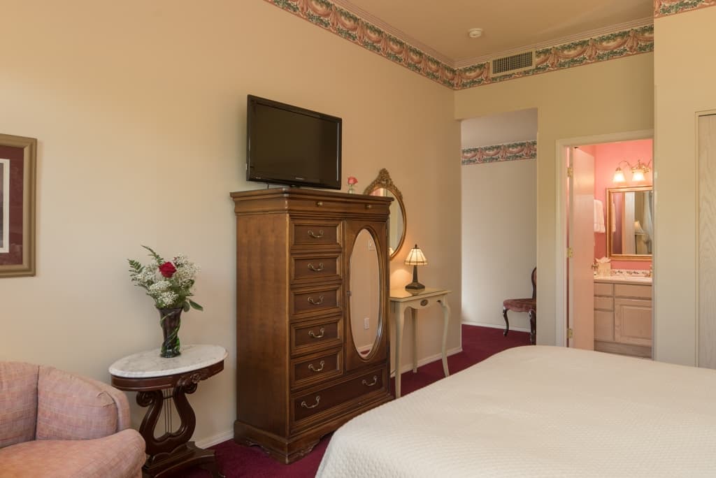 A cozy bedroom featuring a wooden armoire with a TV, a pink armchair, and a side table with flowers. A peek into an adjoining bathroom is visible.