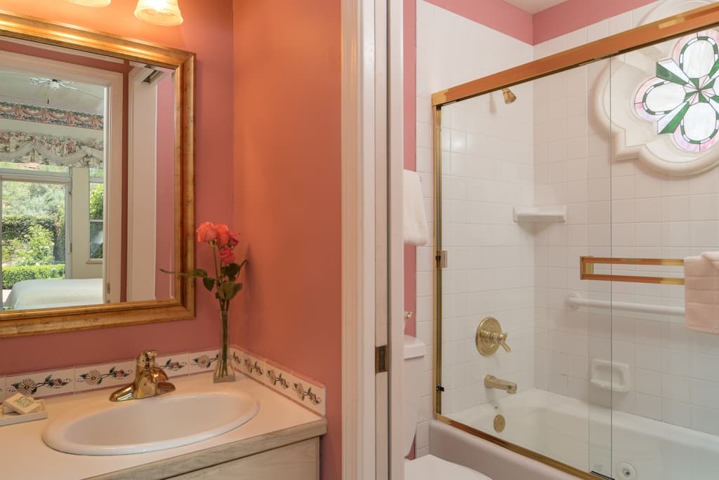 A charming bathroom with pink walls, a vanity with a sink and mirror, and a shower-tub combo with a decorative stained-glass window.
