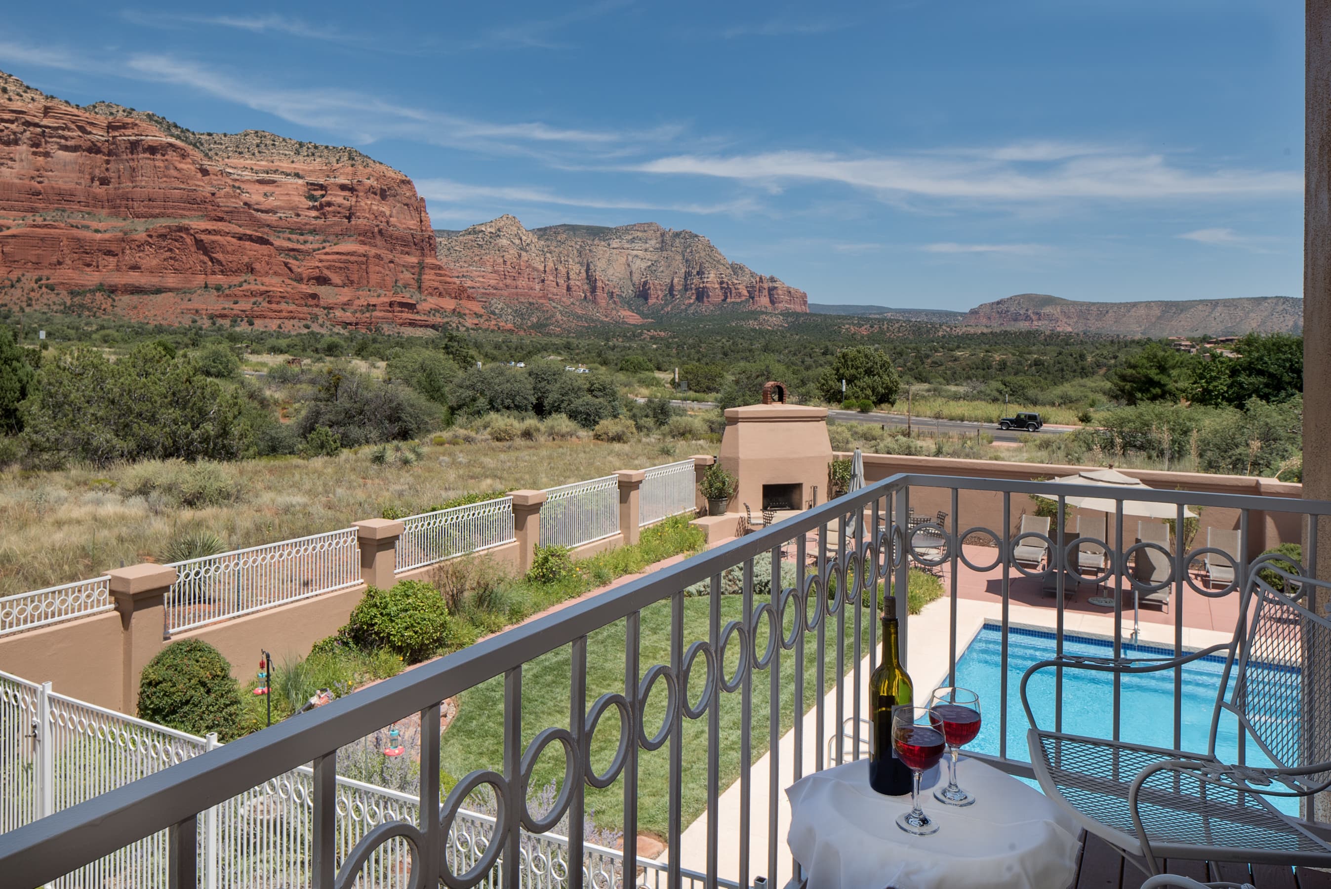 A picturesque balcony with a small table and wine, overlooking a swimming pool, lush landscape, and majestic red rock formations under a clear sky.