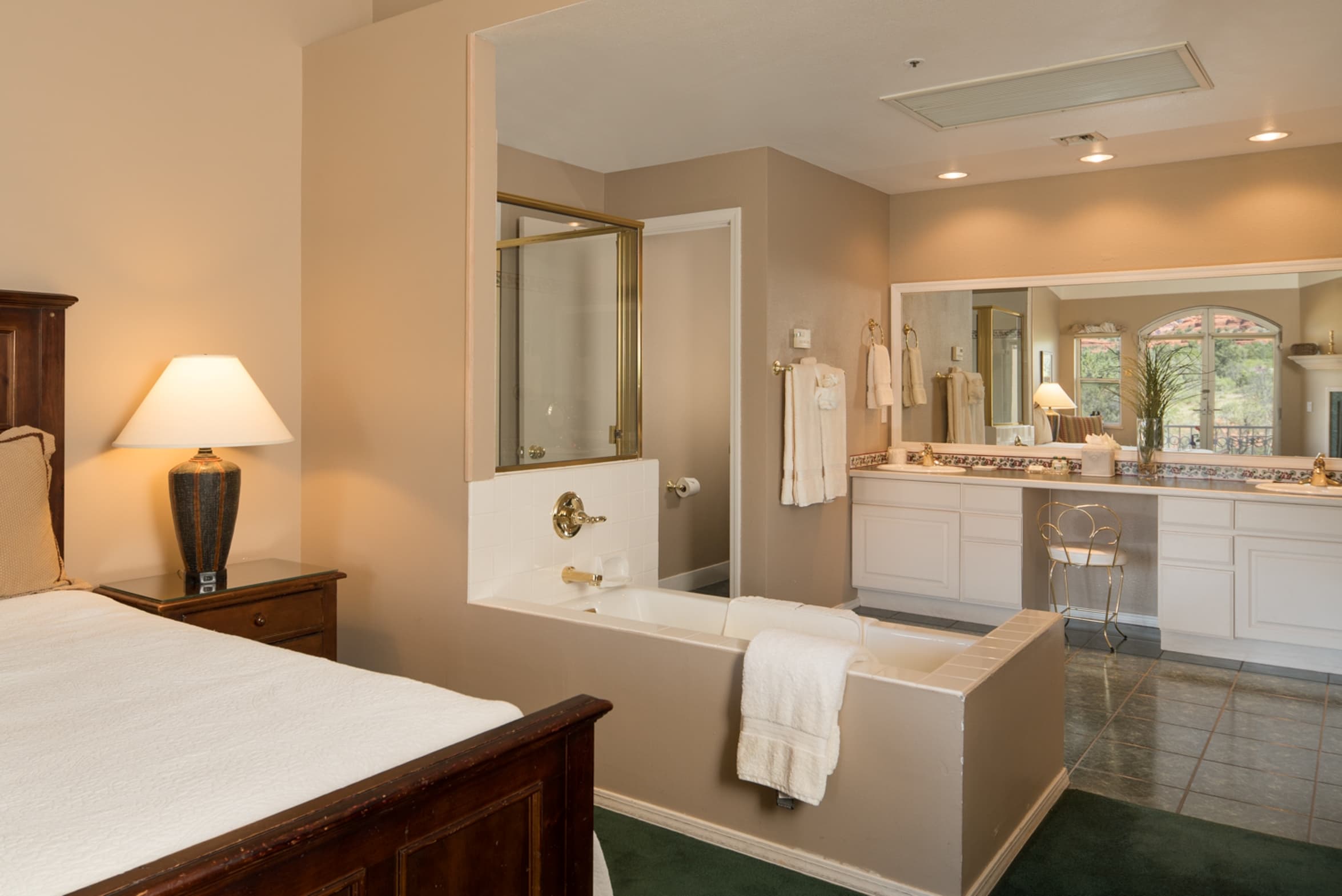 A spacious suite featuring a bedroom area with a king bed and lamp, connected to a luxurious open-concept bathroom with a tub and double vanity.