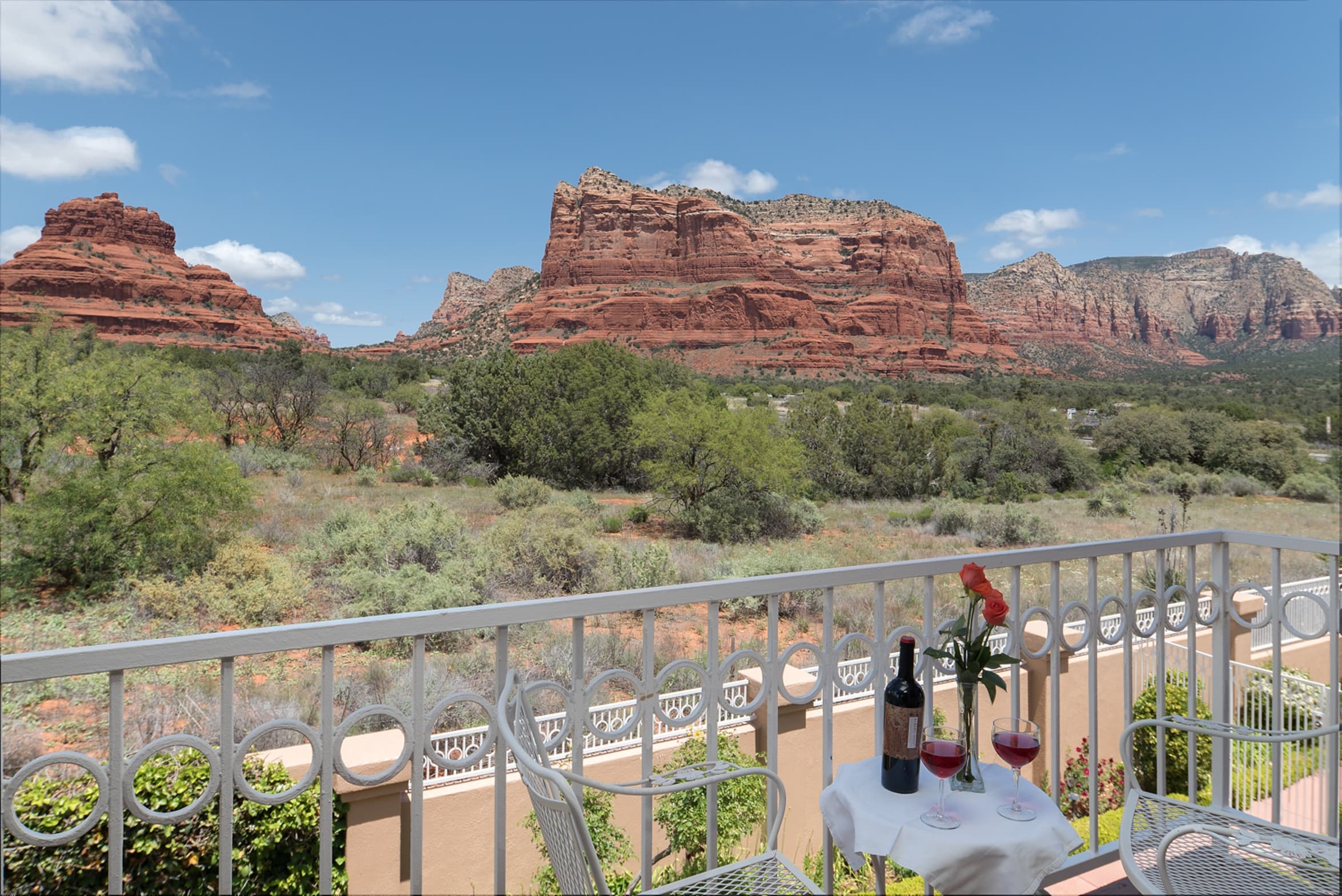 A balcony with a table, wine, and flowers, offering panoramic views of majestic red rock formations and a sprawling landscape under a bright blue sky.