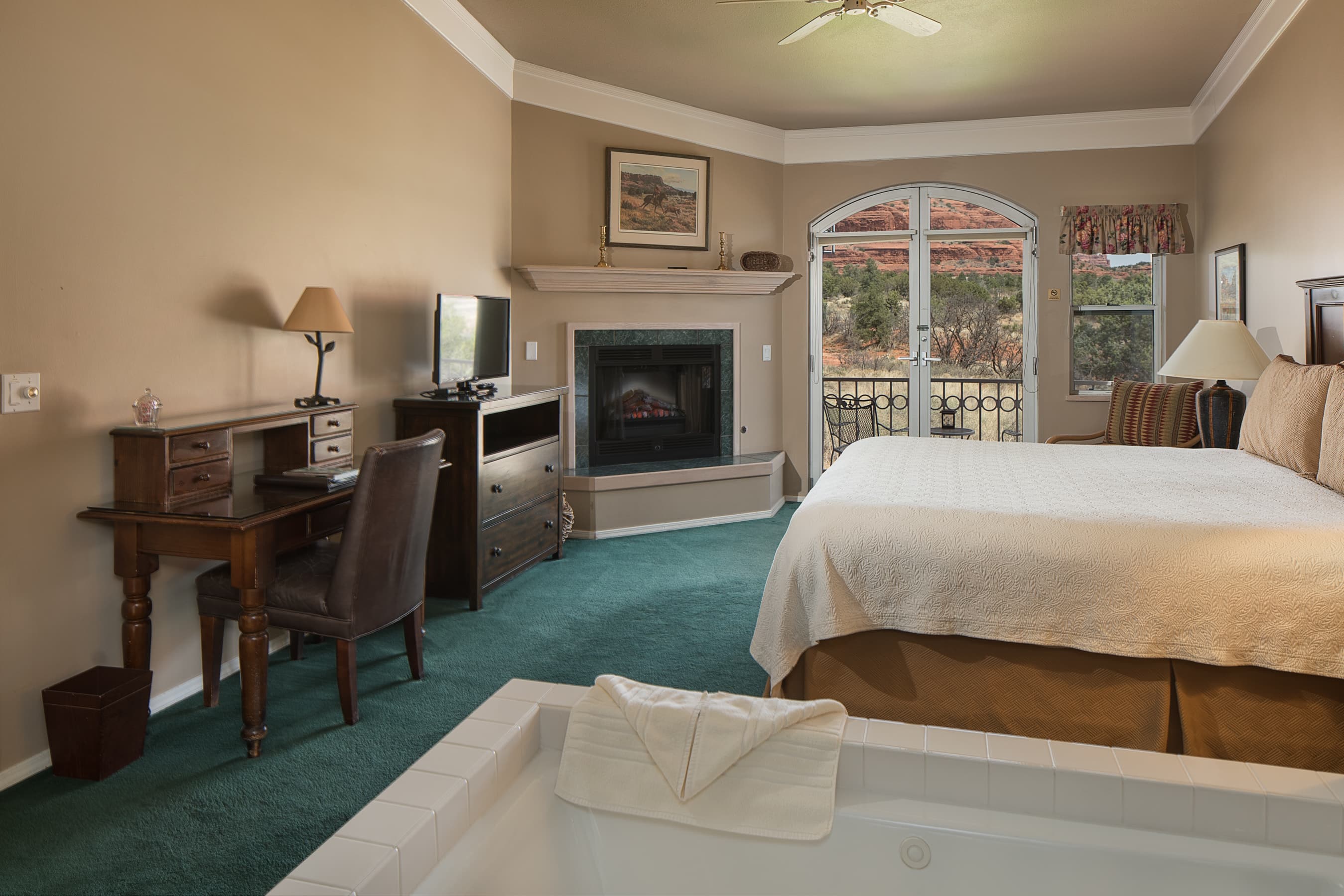 A luxurious room featuring a king bed, fireplace, jetted tub, and a desk, with a balcony offering stunning views of red rock landscapes.