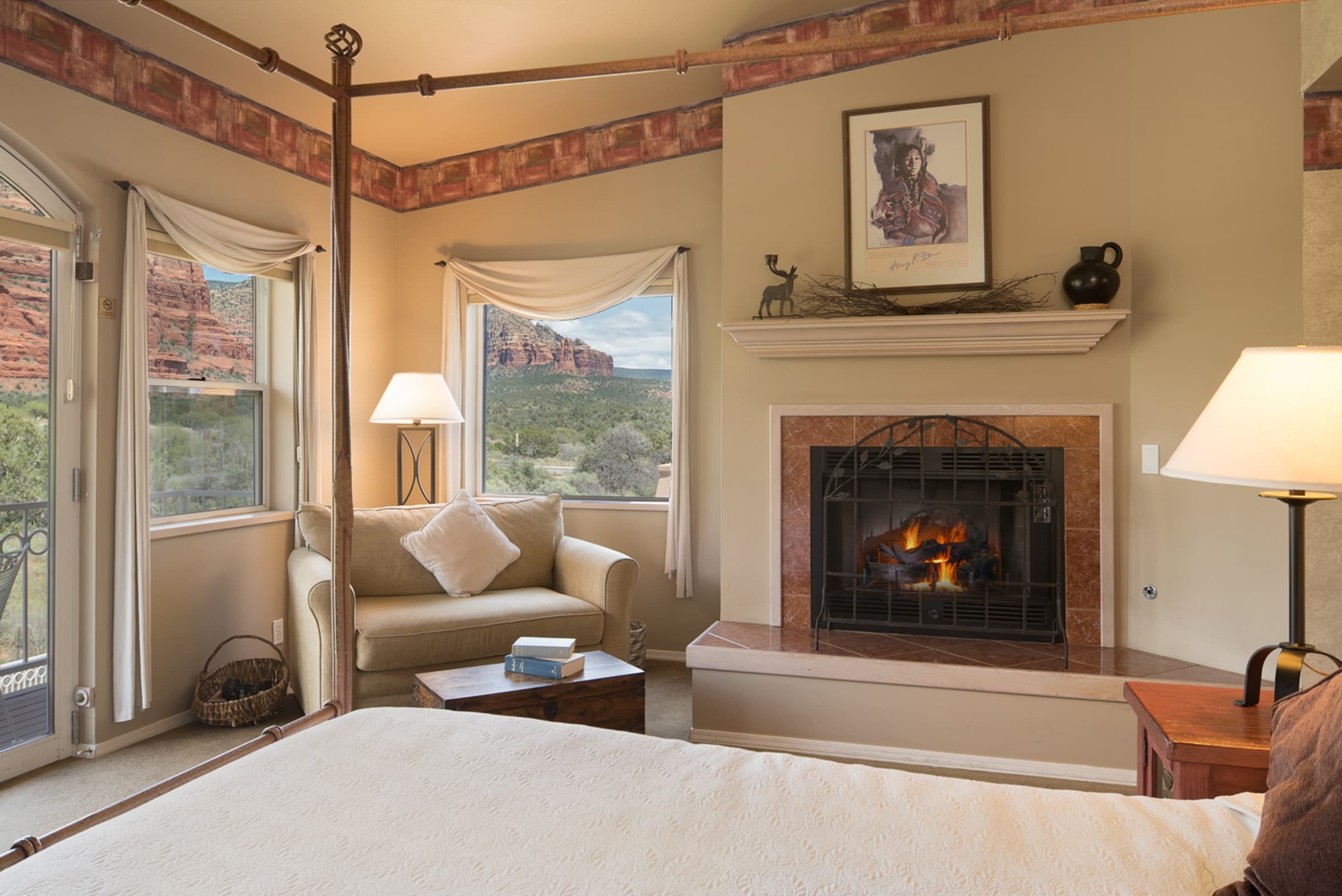A warm and inviting room with a lit fireplace, a cozy loveseat, and a four-poster bed. Large windows and a door offer views of red rock formations.