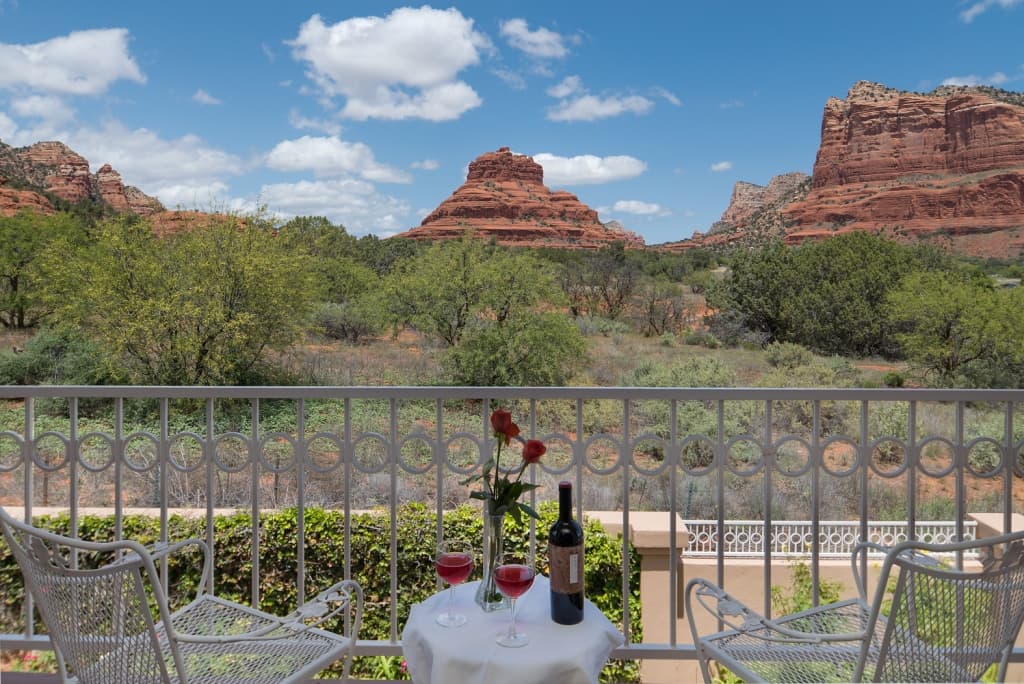 A charming balcony with a table, wine, and roses, offering breathtaking panoramic views of majestic red rock formations and lush greenery under a blue sky.