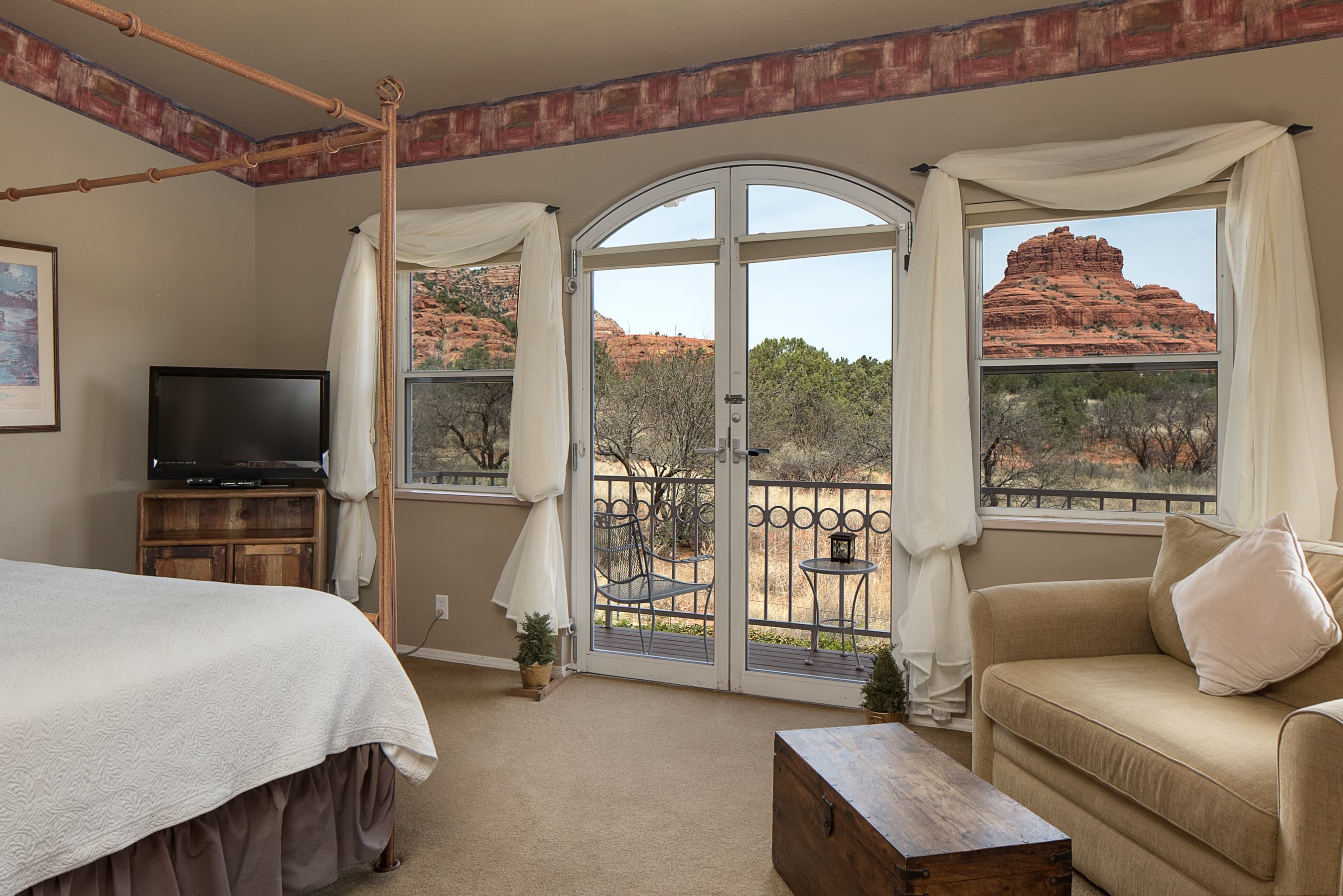 A bright room with a four-poster bed, TV, and a cozy seating area. Arched glass doors lead to a balcony with stunning views of red rock formations.