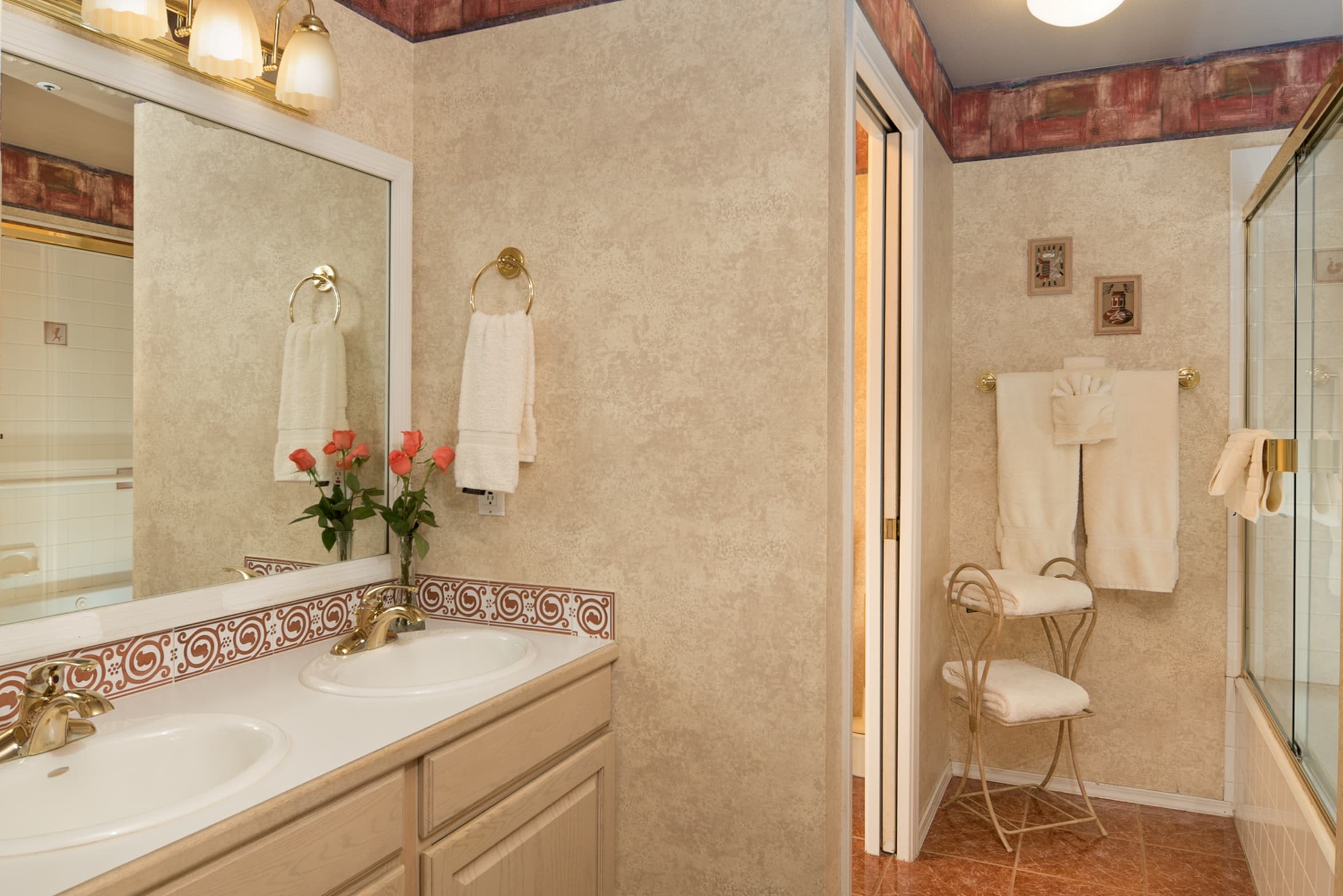 A bathroom with a double vanity, a large mirror, and textured wallpaper. It features warm lighting and a shower/tub area with a chair for convenience.