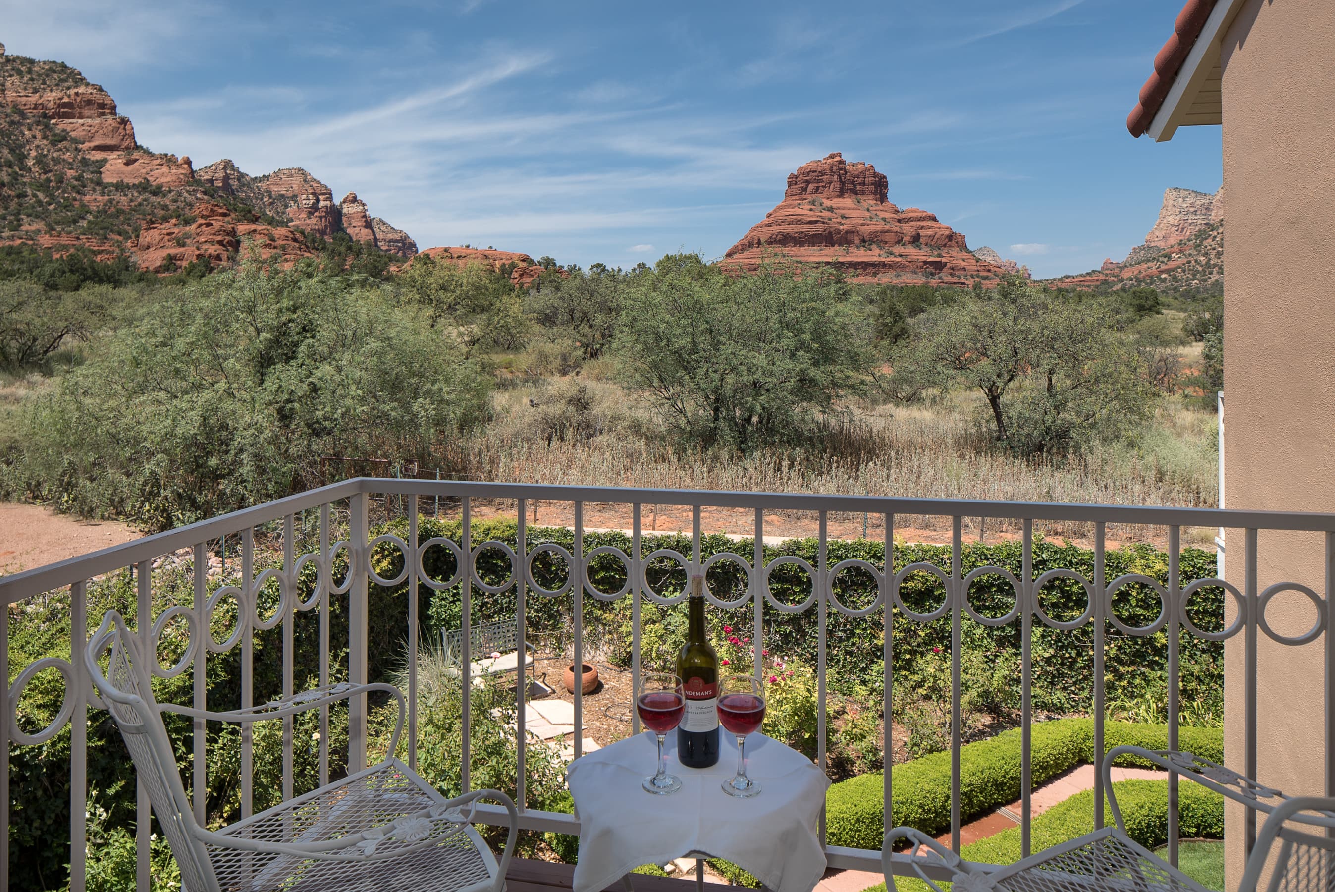 A balcony with a small table and wine, offering stunning panoramic views of vibrant red rock mountains and lush green landscape under a bright blue sky.