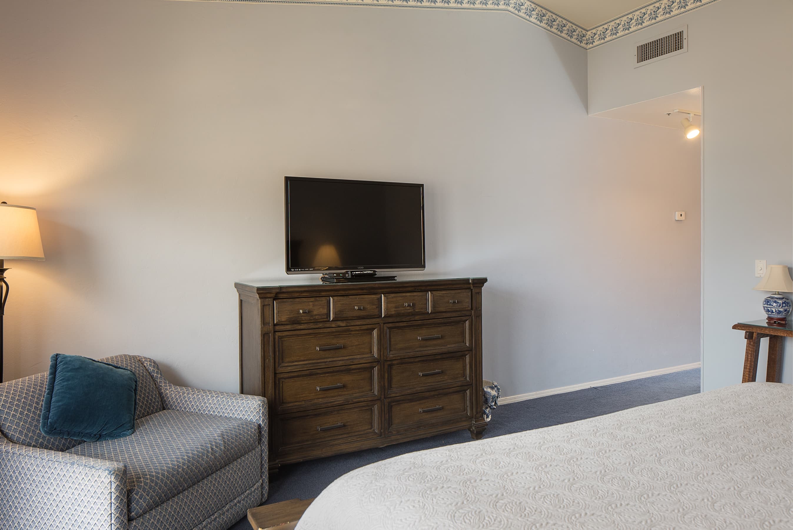 A cozy bedroom featuring a comfortable armchair, a wooden dresser with a TV, and a glimpse of a bed, set against light blue walls.