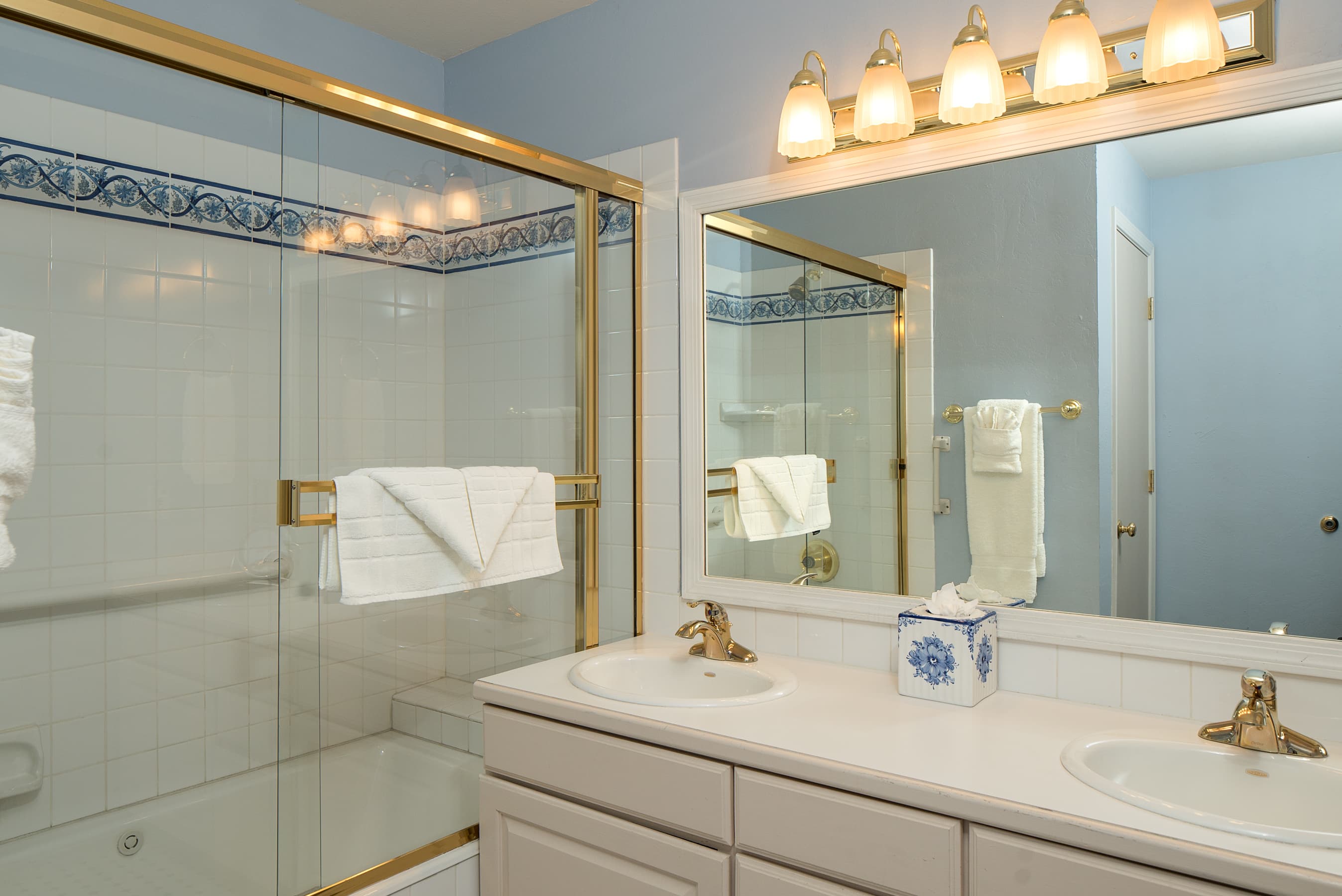 A bathroom with a double vanity, a large mirror, and a shower-tub combo with gold-framed glass doors and blue-and-white patterned tile accents.