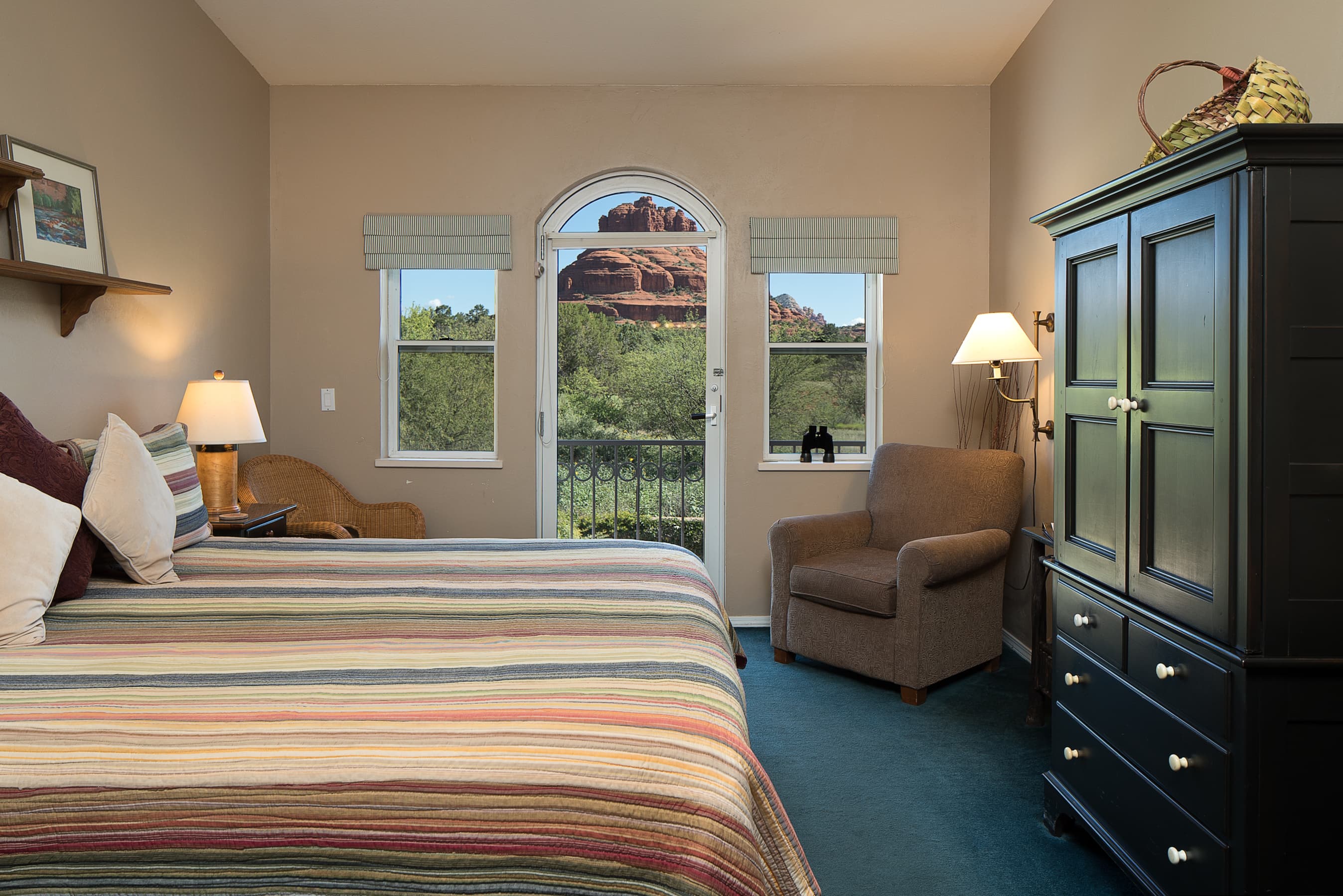 A bedroom with a striped bedspread, a dark wooden armoire, and a cozy armchair. An arched glass door opens to a balcony with views of red rock formations.