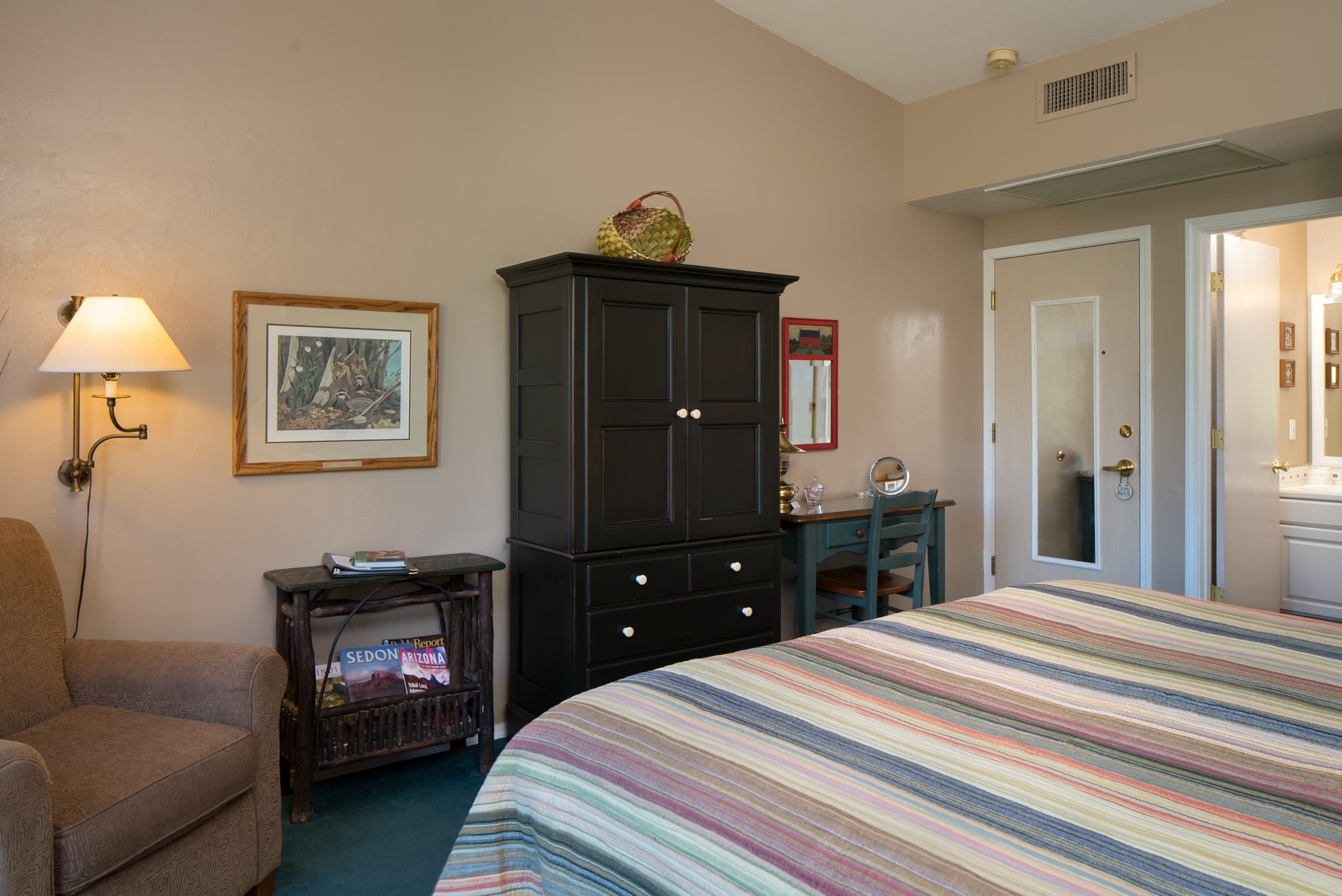 A cozy bedroom featuring a striped bedspread, a dark armoire with a TV, a desk, and an armchair. A doorway leads to an adjacent bathroom.