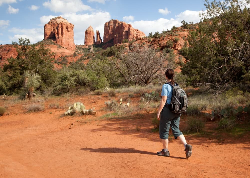 A person hiking on a dusty trail with red rock formations in the background.