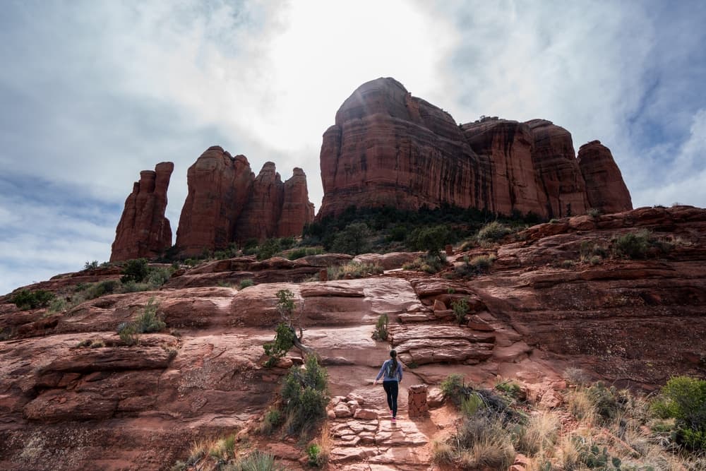A person hiking up rocky terrain beneath towering red rock formations under a cloudy sky.
