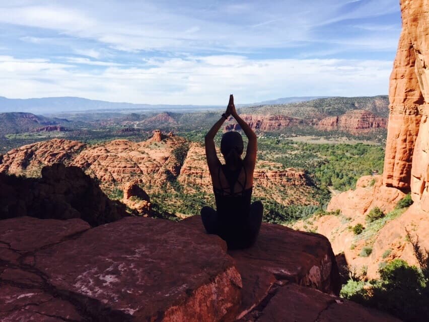 A person sits in a yoga pose on a rocky overlook, surrounded by a vast landscape of red rock formations and greenery under a blue sky.