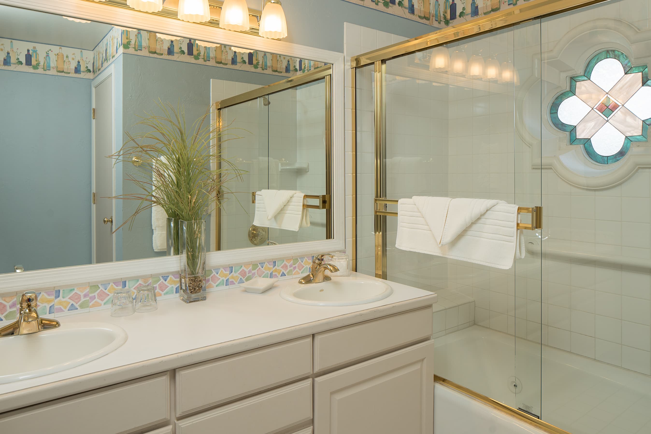 A brightly lit bathroom with a double vanity, a large mirror, and a tub/shower combo with a decorative stained-glass window.