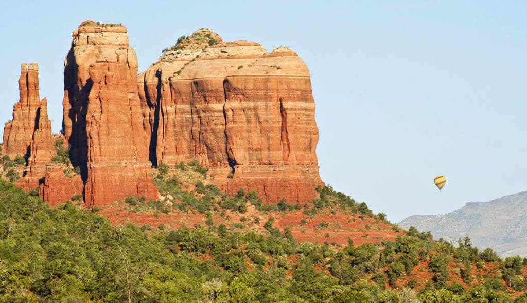 A hot air balloon floats near towering red rock formations against a clear blue sky.