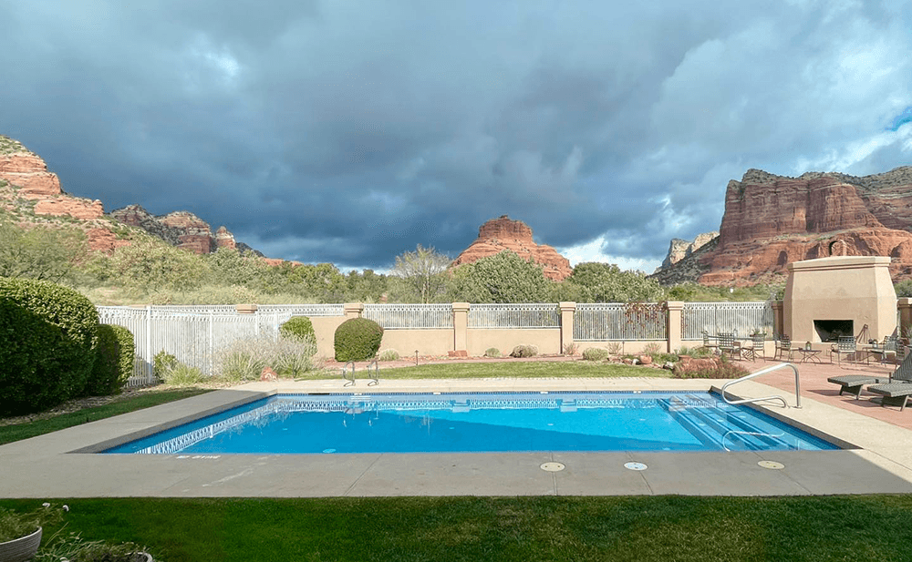 A serene swimming pool surrounded by lush greenery and red rock formations under a cloudy sky.