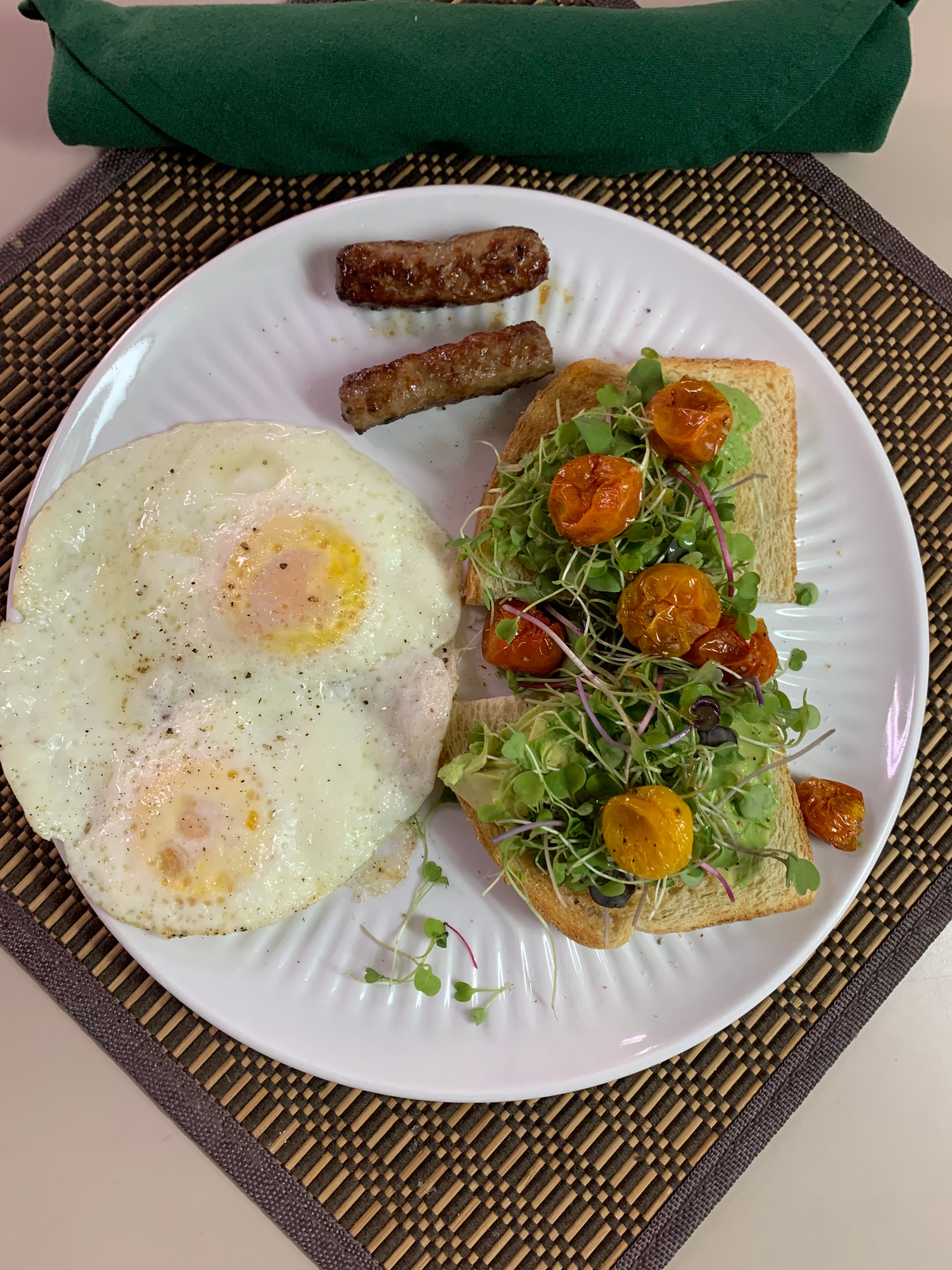 A plate featuring two fried eggs, sausage links, and a slice of toast topped with greens and roasted tomatoes.