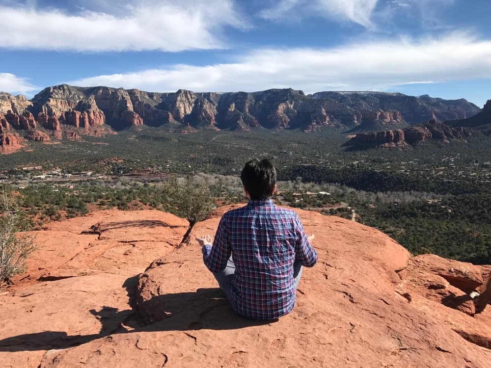 A person sits on a rock ledge, overlooking a vast desert landscape and distant mountains under a blue sky.