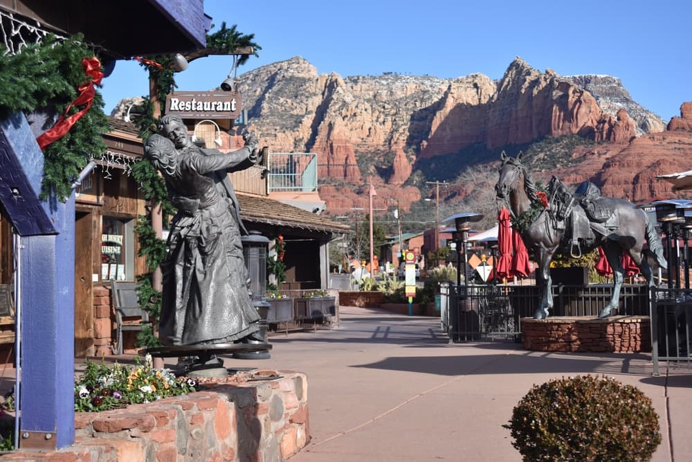 A charming village street features a statue and horse sculpture with red rock mountains in the background.