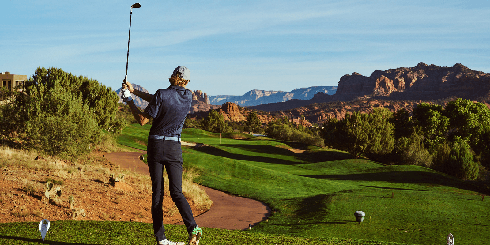 A golfer swings a club on a scenic course with red rock formations in the background.
