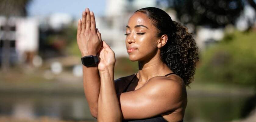 A woman with curly hair practices a yoga pose outdoors by a body of water.