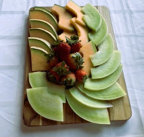 A wooden platter featuring arranged slices of cantaloupe, honeydew, and fresh strawberries.