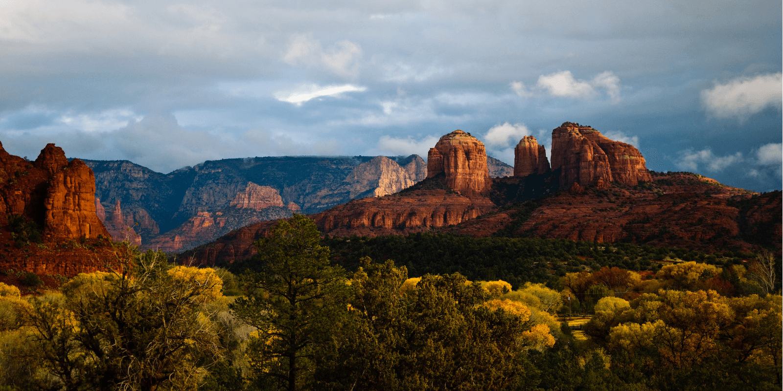 Dramatic red rock formations under a cloudy sky, surrounded by vibrant autumn foliage.