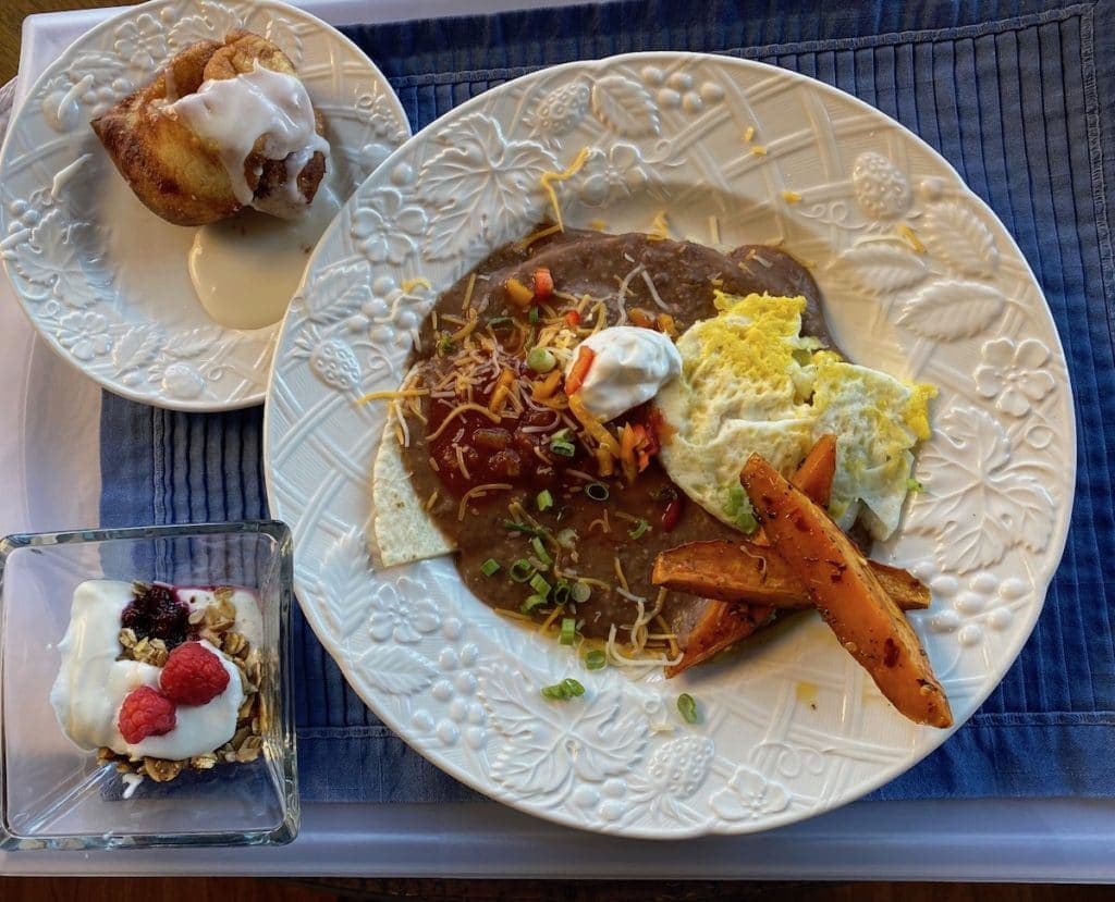 A plate of huevos rancheros with refried beans, topped with cheese and sour cream, accompanied by sweet potato fries, a glass of yogurt with granola and raspberries, and a cinnamon roll.