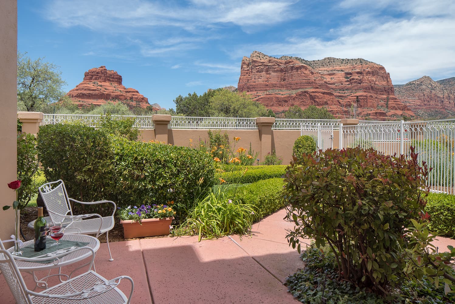 A charming outdoor patio with a table and chairs, surrounded by lush gardens, offering breathtaking views of red rock formations under a blue sky.