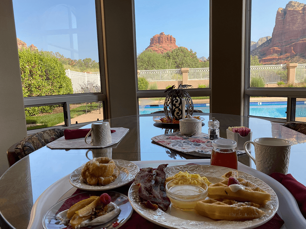 A breakfast table set with various dishes, showcasing a view of red rock formations and a swimming pool through large windows.